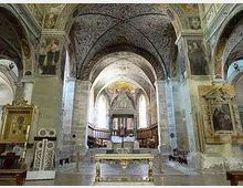 Interno di una chiesa storica con navate in pietra, archi a tutto sesto e volte affrescate; al centro si vede l’altare e, sullo sfondo, l’abside illuminata. Ai lati sono presenti cappelle e monumenti lapidei, con pavimento in lastre e un percorso centrale verso il presbiterio.