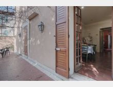 Cortile esterno pavimentato in mattoni lungo la facciata di una casa intonacata, con persiane in legno e lanterne a parete. Una portafinestra aperta collega al soggiorno con tavolo da pranzo e pavimento in cotto; sul fondo si intravede un corridoio interno.