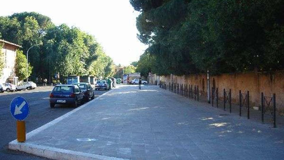 Ampio marciapiede pavimentato lungo una strada urbana con auto parcheggiate a sinistra e una lunga fila di alberi e recinzione metallica sul lato destro. Sullo sfondo si vedono edifici bassi e segnaletica stradale, in un contesto di viale alberato.