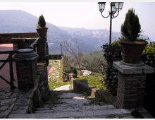 Scalinata in pietra che scende da una terrazza con muretti in muratura e vasi con arbusti, affiancata da un lampione a più luci. Sullo sfondo si vede una valle con uliveti e un borgo collinare in lontananza.