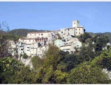 Borgo collinare con case in pietra e intonaco chiaro disposte su un pendio, dominato da un edificio con torre quadrata. Vegetazione fitta e alberi in primo piano, con colline boscose sullo sfondo sotto un cielo sereno.