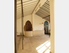 Interno di un ambiente in stile industriale con pareti bianche e soffitto inclinato a pannelli e travi metalliche, sostenuto da sottili colonne scure. Sul lato sinistro c’è una nicchia ad arco con roccia a vista sopra una base in pietra, mentre a destra si vede una porta ad arco vetrata con inferriate.