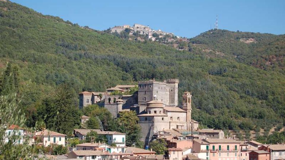 Veduta di un borgo collinare con un grande castello in pietra e una chiesa con campanile, circondati da case dai tetti chiari. Sullo sfondo si estendono pendii boscosi e creste montuose con un altro centro abitato in alto.