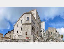 Grande edificio in pietra chiara in stile medievale, con finestre ad arco e una torre laterale, visto dal basso oltre un muro di cinta in pietra. Intorno si notano altri edifici storici e un lampione, sotto un cielo azzurro con nuvole.