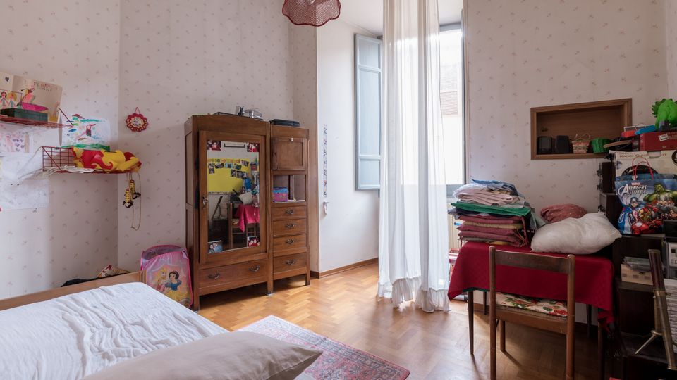 Interno di una camera da letto con pareti rivestite di carta da parati chiara, pavimento in parquet e grande finestra con tende bianche e persiane aperte. Arredi in legno includono un letto, un armadio e un tavolo con sedie, con mensole e oggetti domestici lungo le pareti.