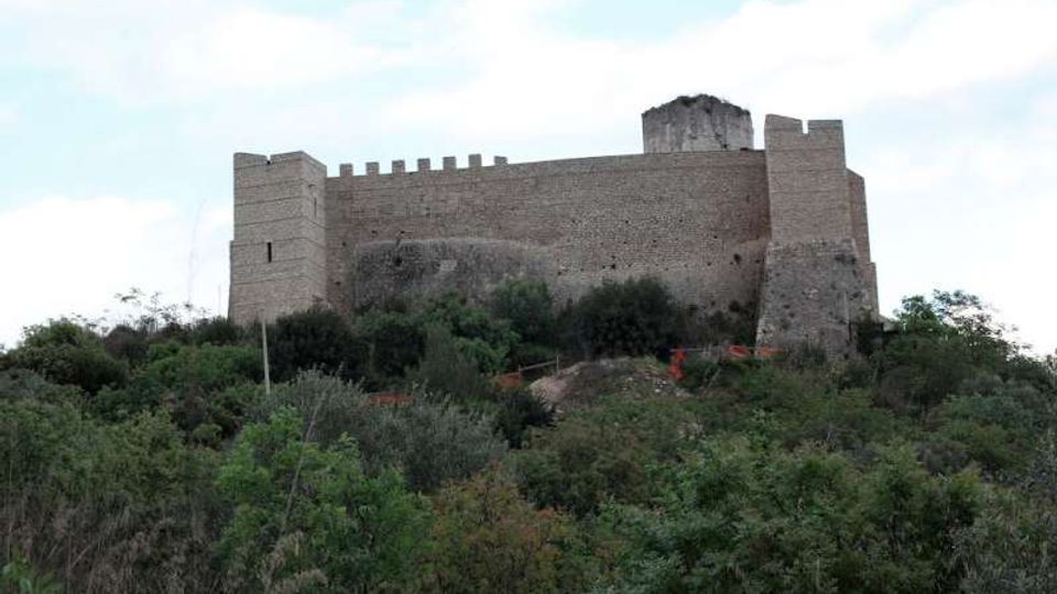 Castello in pietra su una collina, con alte mura merlate e torri angolari. In primo piano vegetazione fitta di macchia mediterranea e arbusti, con cielo chiaro sullo sfondo.