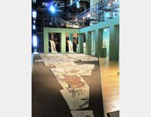 Interno di un museo in un ambiente industriale con passerelle metalliche e tubazioni a vista, dove una lunga pavimentazione a mosaico frammentata è esposta al centro. Strutture verdi con aperture incorniciano l’area espositiva e ospitano statue su piedistalli lungo il percorso.