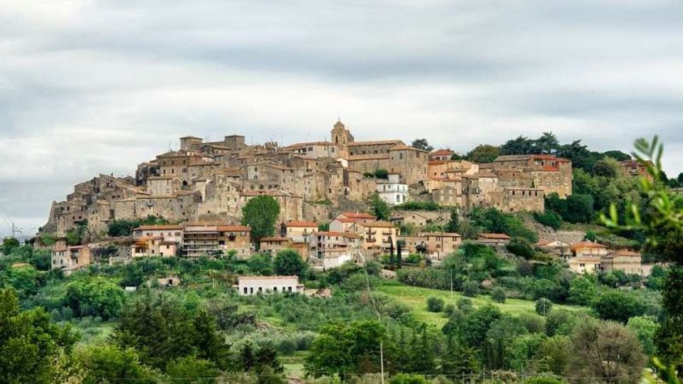Borgo collinare con case in pietra addossate tra loro e una chiesa con campanile in cima, circondato da vegetazione. In primo piano si vede una valle verde con alberi e campi sotto un cielo nuvoloso.