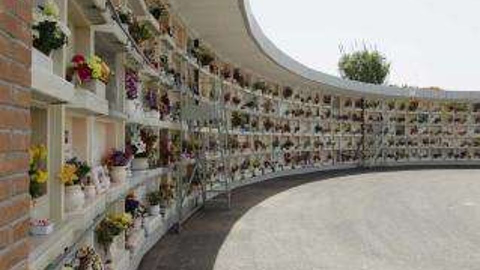 Viale curvo di un cimitero con file di loculi bianchi lungo il lato sinistro, decorati con fiori e targhe, sotto una copertura continua. Pavimentazione in cemento e cielo grigio sullo sfondo.