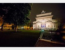 Veduta notturna di un edificio monumentale in stile neoclassico, illuminato, con grande facciata simmetrica e frontone decorato, affacciato su un giardino pubblico. In primo piano si vede un percorso con parapetto in vetro, alberi e prato che circondano la piazza.