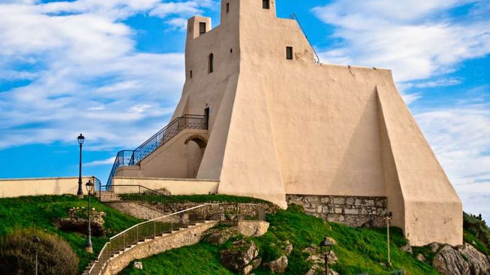 Fortezza in muratura chiara su un promontorio erboso e roccioso, con pareti inclinate, piccole finestre e una torre superiore. Una scalinata con ringhiere e lampioni conduce all’ingresso, sotto un cielo azzurro con nuvole.