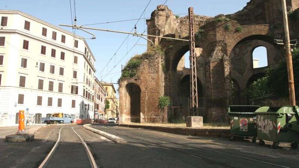 Strada urbana con binari del tram e cavi aerei, affiancata da un grande muro in mattoni con arcate in rovina. Sullo sfondo si vede un edificio bianco a più piani e l’area stradale con marciapiedi e cassonetti.