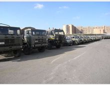 Parcheggio asfaltato all’aperto con una lunga fila di camion e veicoli militari verde oliva allineati. Sullo sfondo si vedono grandi edifici in cemento e mattoni sotto un cielo sereno.