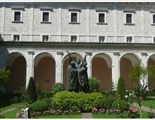 Cortile interno con giardino curato e aiuole fiorite, al centro una statua in bronzo su basamento. Sullo sfondo si vede un edificio in pietra con porticato ad archi e finestre regolari ai piani superiori.