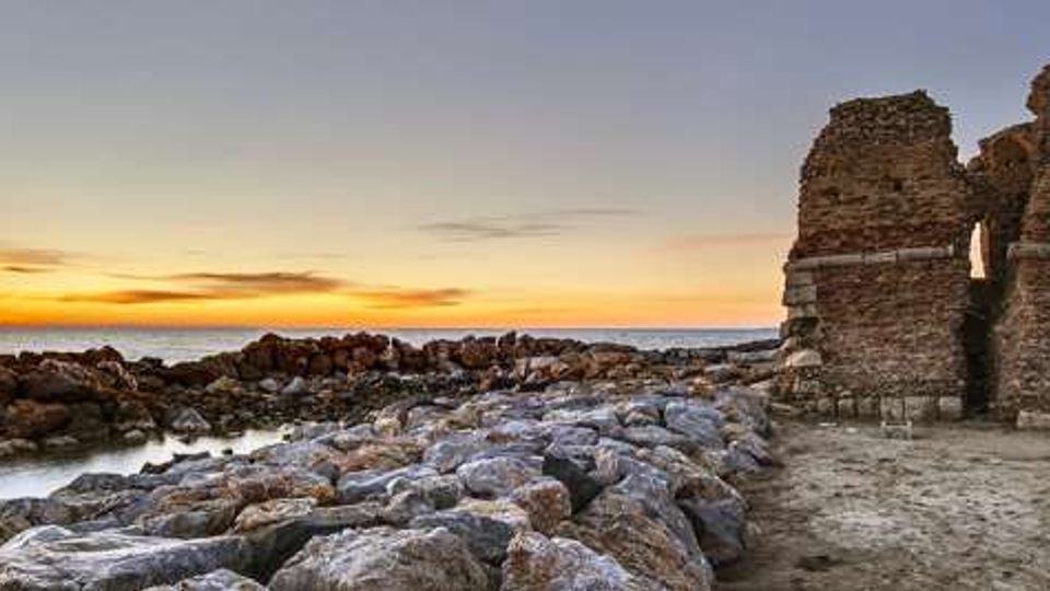 Rovine in pietra di un’antica struttura costiera con due muri parzialmente crollati e aperture, su una spiaggia rocciosa. In primo piano una scogliera di massi conduce al mare calmo, con il sole al tramonto all’orizzonte.