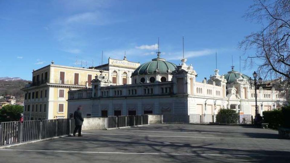 Edificio monumentale in stile classico con facciata chiara, finestre ad arco e grandi cupole verdi, visibile oltre una piazza asfaltata con ringhiere e lampioni. Sullo sfondo si notano colline e un cielo azzurro limpido.