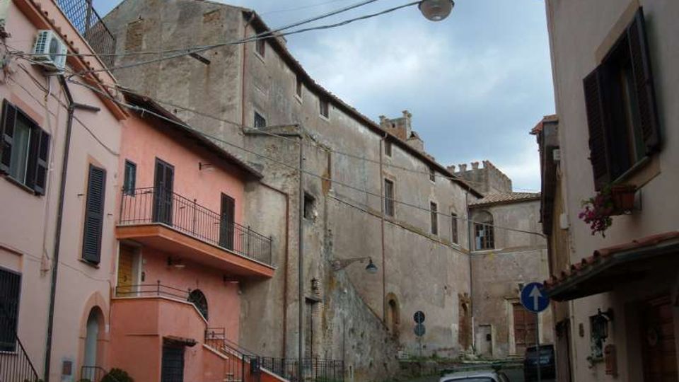 Stradina in pietra in un borgo storico, fiancheggiata da edifici in intonaco rosa e grigio con persiane, balconi e cavi aerei. Sullo sfondo si vede una struttura in muratura dall’aspetto medievale e un lampione sospeso sopra la via.