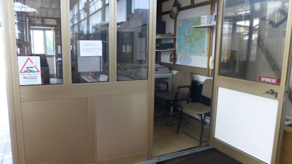 Ingresso di un piccolo ufficio o postazione di controllo con pareti vetrate e struttura metallica, porta aperta con cartello “SPINGERE”. All’interno si vedono sedie, una scrivania con attrezzature e una mappa appesa alla parete.