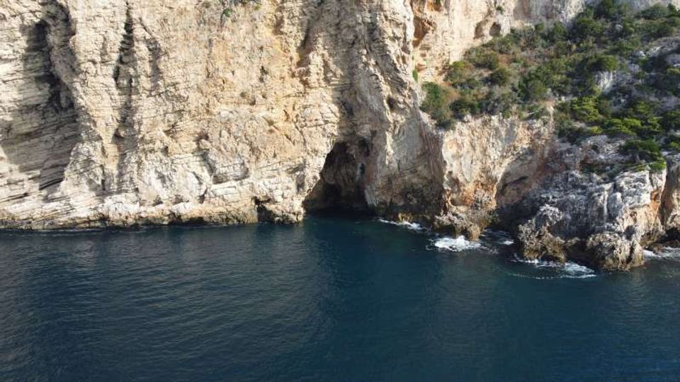 Scogliera costiera di roccia chiara con una piccola grotta alla base, affacciata su mare blu scuro. Sul promontorio sono visibili macchia mediterranea e massi frastagliati con onde che si infrangono vicino alla riva.