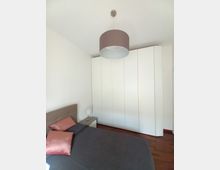 Camera da letto moderna con letto matrimoniale in primo piano, comodino con lampada e grande armadio bianco a pannelli lungo la parete. Soffitto bianco con lampadario a sospensione cilindrico e pavimento in legno scuro.
