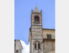 Campanile in pietra e mattoni con cuspide piramidale, caratterizzato da aperture ad arco e una cella campanaria con ringhiera. La torre è addossata a edifici in muratura in un contesto di borgo, sullo sfondo di cielo azzurro.