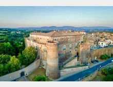 Veduta aerea di un grande castello in pietra con mura merlate e torri cilindriche, circondato da una cinta muraria e affacciato su una strada. Sullo sfondo si vede un borgo con edifici in pietra e un paesaggio collinare verde fino alle montagne all’orizzonte.