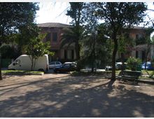 Piccolo parco urbano con alberi alti che proiettano ombre su un’area in terra battuta e una panchina verde. Sullo sfondo si vedono auto parcheggiate e un edificio in mattoni a due piani con tetto in tegole, parzialmente nascosto dalla vegetazione.