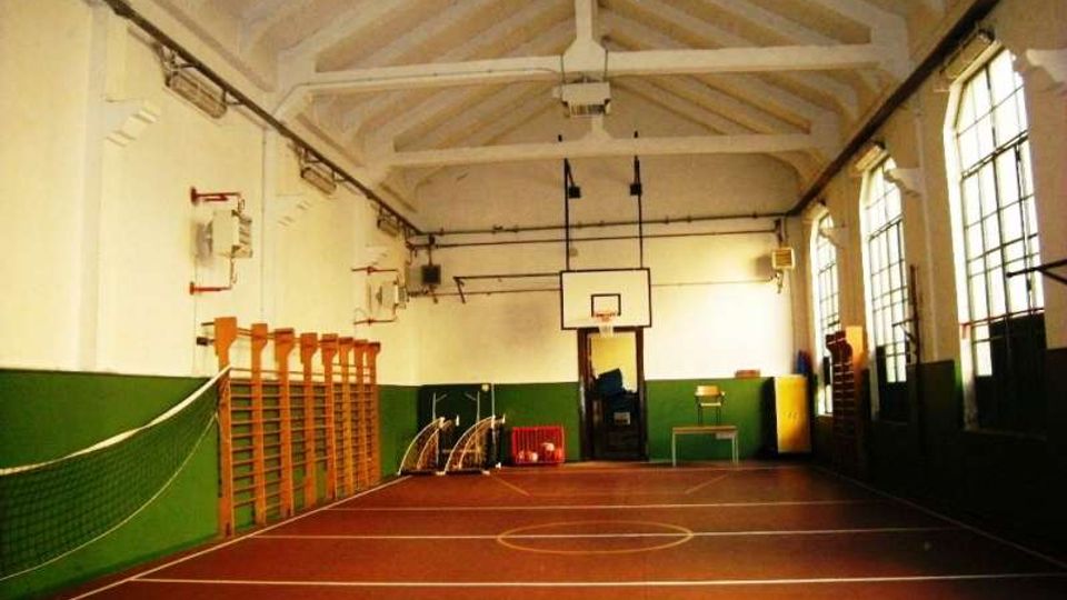 Palestra scolastica al coperto con campo segnato a terra e canestro da basket su una parete di fondo. Soffitto alto con travi a vista, pareti bianche e verdi, spalliere in legno e grandi finestre laterali.