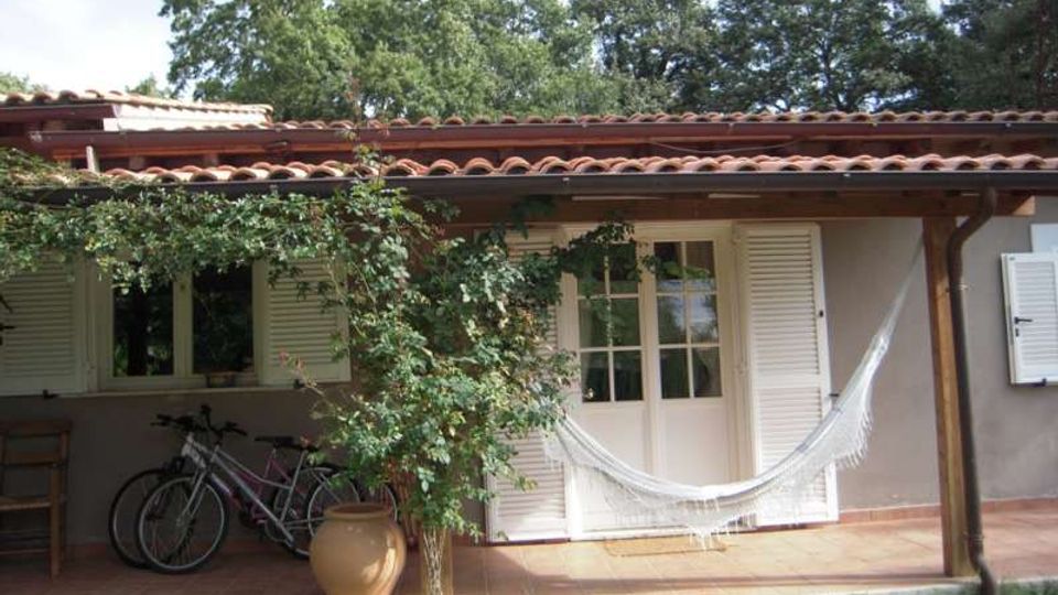 Veranda di una casa a un piano con tetto in tegole e colonne in legno, pavimento in cotto e porte-finestre con persiane bianche. Davanti c’è un’amaca appesa, una pianta rampicante in vaso e biciclette sotto il portico, con prato e alberi sullo sfondo.