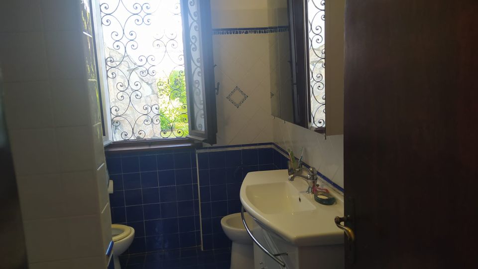 Piccolo bagno con piastrelle blu e bianche, lavabo con mobile e specchio sulla parete. Una finestra aperta con inferriata decorativa lascia entrare luce naturale, con vegetazione visibile all’esterno.