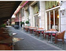 Dehors di un ristorante lungo un portico coperto, con file di tavoli e sedie in rattan su pavimentazione in pietra. Sul lato destro si vede la facciata in pietra con grandi vetrate e tende, illuminate da applique a parete.