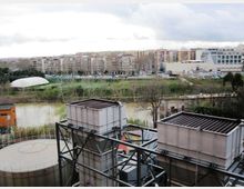 Vista urbana con un fiume in primo piano e, davanti, grandi strutture industriali metalliche e un serbatoio circolare. Sullo sfondo si vedono aree verdi, una strada e palazzi residenziali e commerciali sotto un cielo nuvoloso.