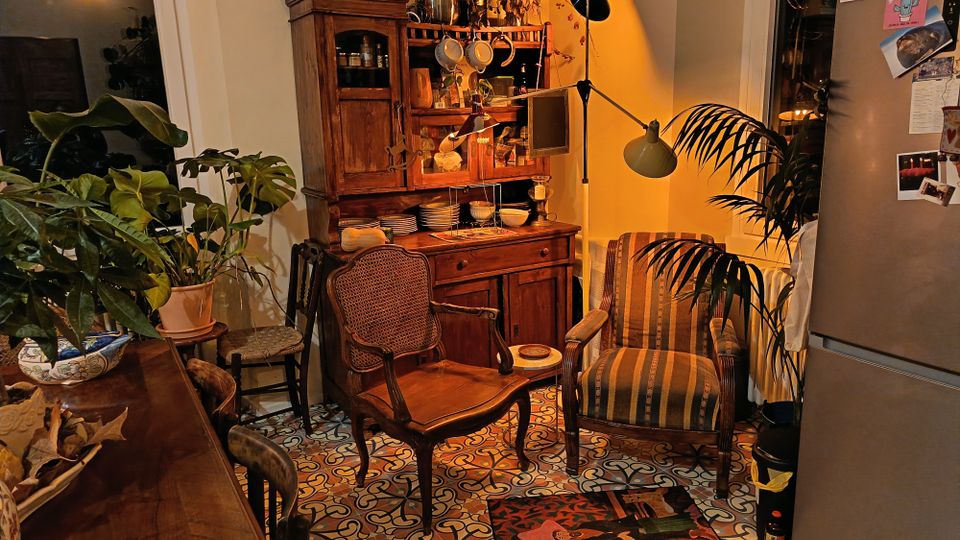 Angolo cucina-soggiorno con mobili in legno massello e credenza piena di stoviglie, lampade da terra e poltrone imbottite. Pavimento con piastrelle decorative a motivi geometrici, molte piante in vaso e frigorifero a vista sulla destra.