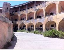 Cortile interno di un edificio storico a pianta semicircolare, con due livelli di logge ad archi e pareti intonacate e scrostate. In primo piano un ampio percorso in pietra affiancato da vegetazione spontanea, con una torre laterale sullo sfondo.