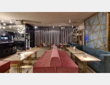Interno di un lounge bar moderno con lunga panca centrale rivestita in tessuto rosso e tavolini bassi in marmo con basi metalliche. Lungo la parete destra ci sono divanetti blu e una parete decorativa con pannelli illuminati, mentre sul fondo si vedono tende grigie e illuminazione a faretti sul soffitto.