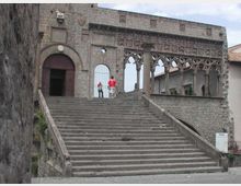 Ampia scalinata in pietra che conduce all’ingresso ad arco di un edificio storico fortificato, con muri in blocchi di pietra. Sul lato destro si estende una loggia con archi gotici traforati affacciata su un cortile interno.