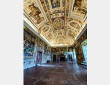 Interno di una grande sala storica con pavimento in pietra, pareti rivestite da ampi affreschi e un grande camino sul fondo. Il soffitto a volta è riccamente decorato con riquadri dipinti, motivi ornamentali e scene figurative.