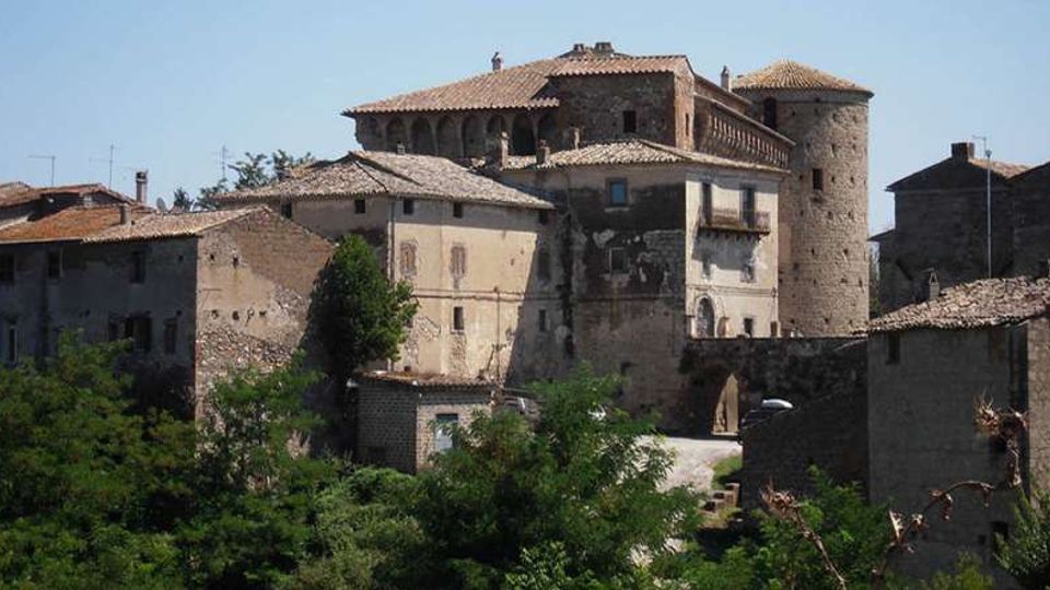 Complesso di edifici storici in pietra con tetti in coppi, collegati tra loro e affiancati da una torre cilindrica tipo fortilizio. In primo piano ci sono alberi e vegetazione, con un piccolo passaggio ad arco tra le costruzioni sotto un cielo limpido.