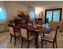 Soggiorno con zona pranzo: grande tavolo in legno scuro con sedie imbottite e centrotavola floreale, su pavimento in marmo chiaro. Sullo sfondo ci sono finestre con tende leggere, una credenza, un quadro, piante e una porta finestra con vetrate rigate accanto a un divano.