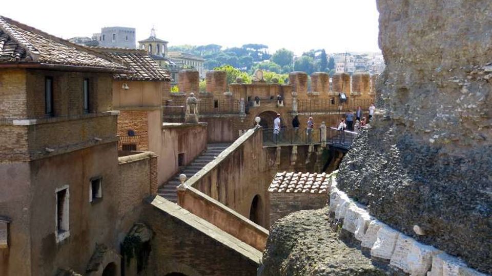 Cortile e passaggi sopraelevati di una fortificazione storica in mattoni, con tetti in tegole e mura merlate. Un camminamento con parapetti e scalinate attraversa la struttura, con la città e colline alberate sullo sfondo.