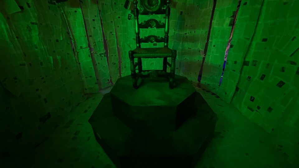 Stanza immersiva illuminata di verde, con pareti e soffitto rivestiti da fogli di carta disposti in modo irregolare. Al centro, una sedia in legno intagliato è collocata su una pedana ottagonale scura.
