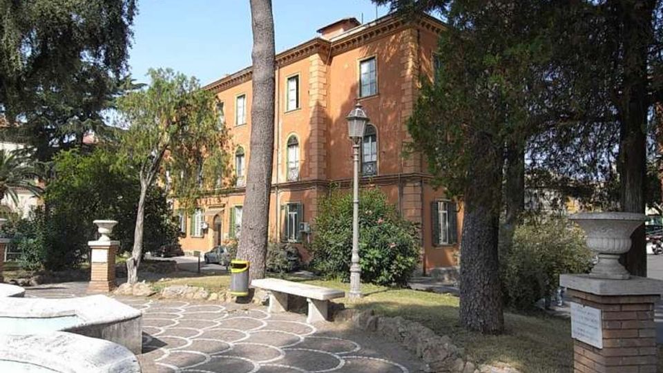 Edificio storico a tre piani con facciata color terracotta e finestre ad arco al piano terra, immerso in un giardino con alti alberi. In primo piano si vede un vialetto pavimentato con motivo a semicerchi, una panchina in pietra e un lampione, con muretti e vasi decorativi ai lati.