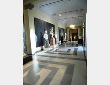 Corridoio interno di un museo o galleria con pavimento geometrico in marmo grigio e bianco e soffitto alto con illuminazione a binari. Lungo le pareti sono esposte statue e busti su piedistalli, con grandi pannelli scuri e un passaggio ad arco sullo sfondo.