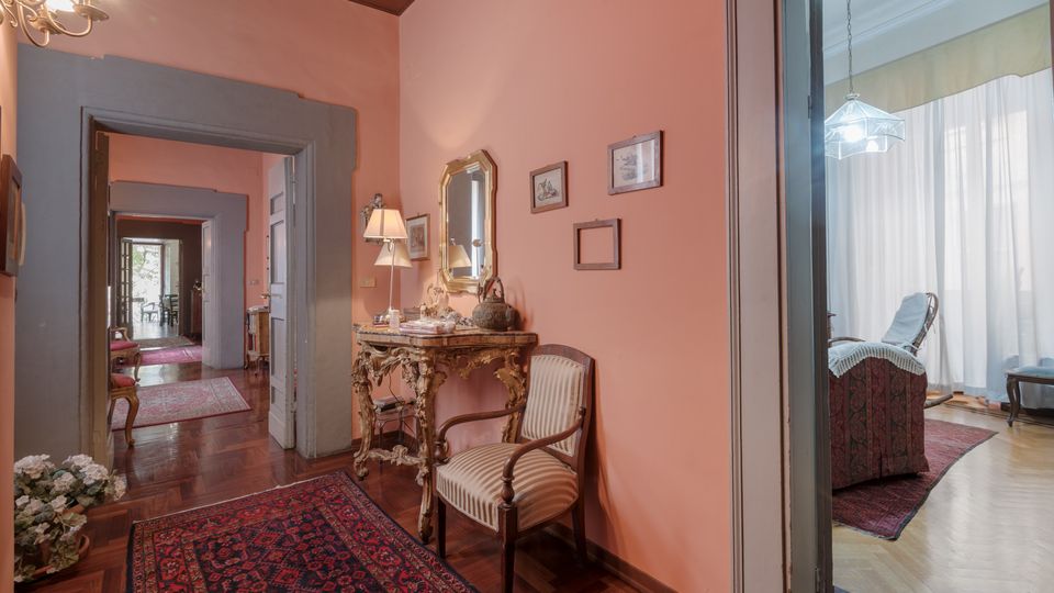 Corridoio interno di una casa con pareti color salmone, pavimento in parquet e tappeto persiano; lungo la parete c’è una consolle decorata con specchio, lampade e quadretti. Porte con cornici grigie si aprono su altre stanze, tra cui un soggiorno luminoso con grandi tende e una lampada a sospensione.