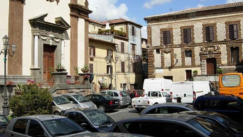 Piazza cittadina con parcheggio, circondata da edifici storici in pietra con persiane e facciate a più piani. A sinistra si vede una chiesa con portale monumentale e orologio sulla facciata, con lampioni e aiuole lungo il bordo della piazza.