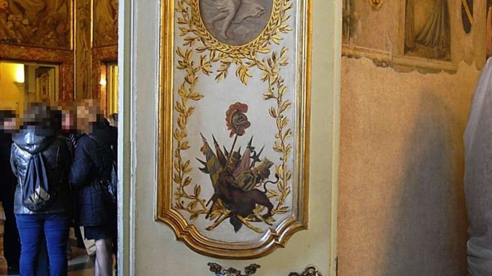 Porta interna decorata in stile barocco, dipinta con medaglioni, ghirlande dorate e motivi araldici, che si apre su una sala storica con lampadario e pareti affrescate. Sullo sfondo si intravedono pavimento a scacchi e ambienti riccamente ornati.