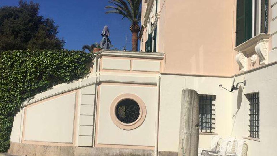 Facciata esterna di un edificio in stile classico color crema e bianco, con cornici decorative, persiane verdi e una finestra circolare in un muretto laterale. In primo piano c’è un prato con ombre di palme; a lato si vedono una panchina metallica e finestre con inferriate.