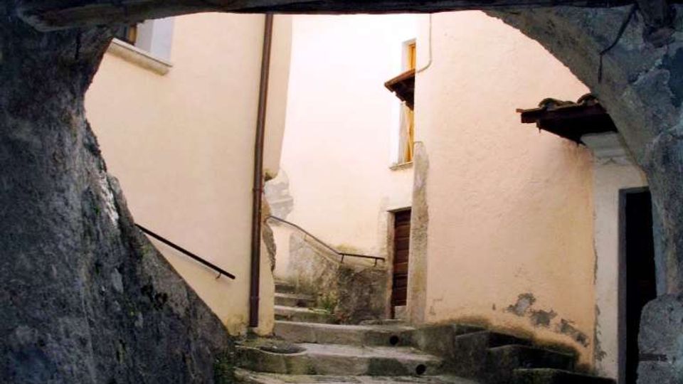 Vicolo stretto in un borgo storico, con gradini in pietra e pavimentazione irregolare che salgono tra muri in intonaco chiaro. La scena è incorniciata da un arco basso con travi di legno scure, con una ringhiera metallica e piccole porte e finestre sulle facciate.