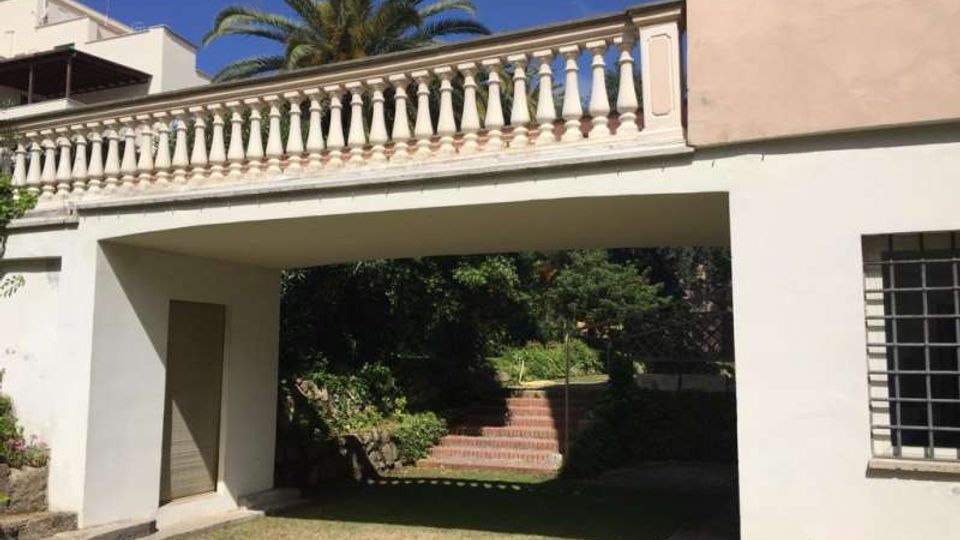 Cortile di una casa con prato e un portico passante sotto una terrazza con balaustra bianca, affiancato da pareti chiare e una finestra con grata. Sullo sfondo si vedono una scala in mattoni che sale tra vegetazione e una palma alta.