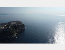 Veduta aerea di una città fortificata su un promontorio roccioso a picco sul mare, con mura e edifici compatti lungo la scogliera. Ampia distesa d’acqua calma con riflessi del sole e costa lontana appena visibile all’orizzonte.